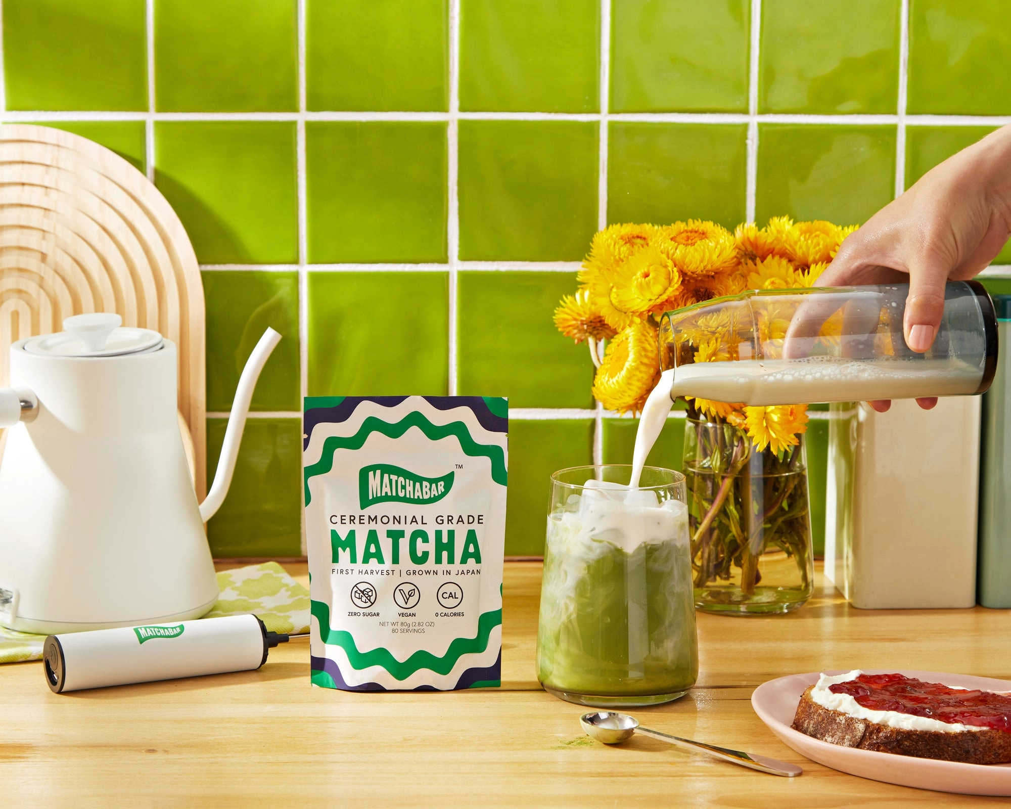 A kitchen counter with a green tile wall, a pouch of matcha, a kettle and a glass of iced matcha being poured