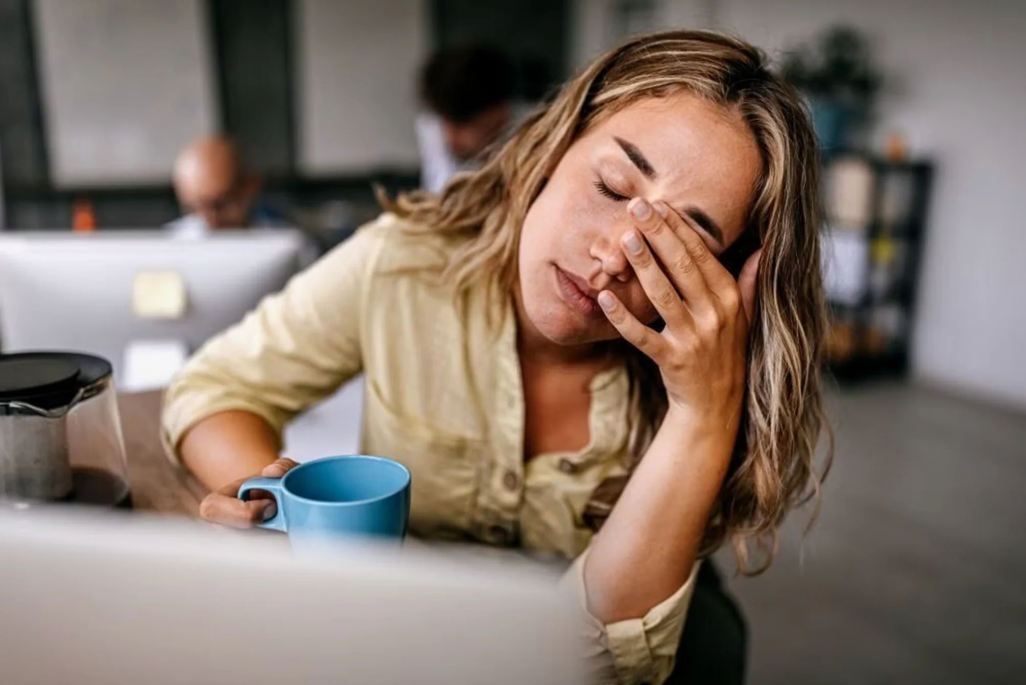 A tired woman at a desk rubbing her eyes in front of a laptop